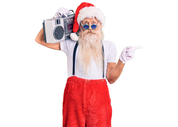 Oude senior man met grijs haar en lange baard in kerstman kostuum en boombox lachend vrolijk wijzend met hand en vinger naar de zijkant  - Foto, afbeelding