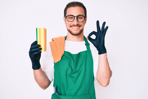 Young handsome man wearing apron holding scourer doing ok sign with fingers, smiling friendly gesturing excellent symbol  - Photo, Image