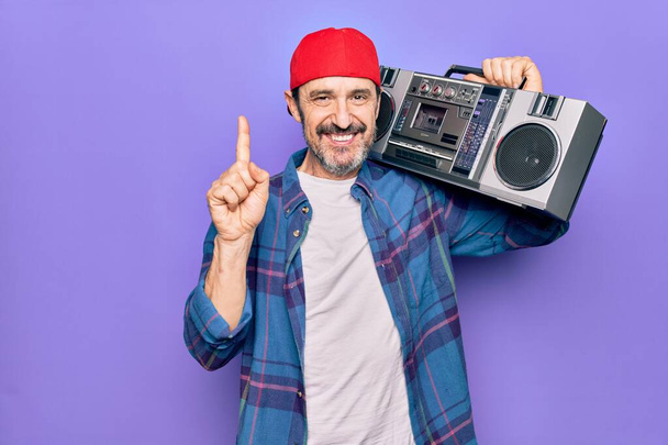Middle age man wearing cap listening vintage boombox over isolated purple background smiling with an idea or question pointing finger with happy face, number one - Φωτογραφία, εικόνα