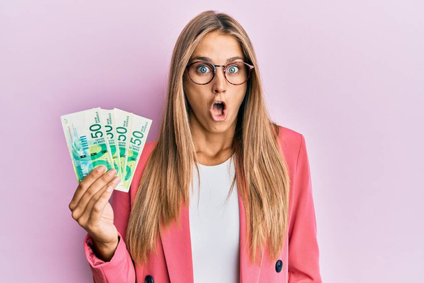 Young blonde woman wearing business style holding israel shekels scared and amazed with open mouth for surprise, disbelief face  - Foto, afbeelding