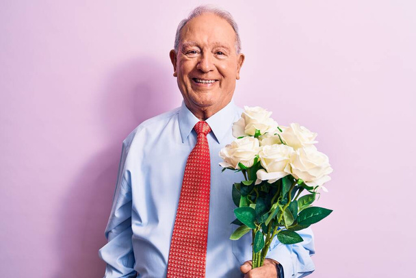Senior hombre de negocios de pelo gris con corbata sosteniendo ramo de flores sobre fondo rosa mirando positiva y feliz de pie y sonriendo con una sonrisa confiada mostrando los dientes - Foto, imagen