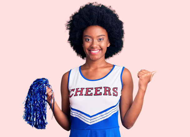 Joven mujer afroamericana vistiendo uniforme de porrista sosteniendo pompón apuntando a la espalda con la mano y los pulgares hacia arriba, sonriendo confiado  - Foto, Imagen