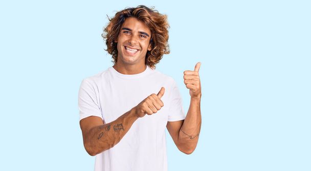 Jeune homme hispanique portant un t-shirt blanc décontracté pointant vers le dos derrière avec la main et les pouces levés, souriant confiant  - Photo, image