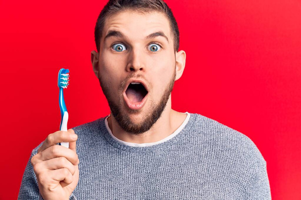 Young handsome man holding toothbrush scared and amazed with open mouth for surprise, disbelief face  - Foto, imagen