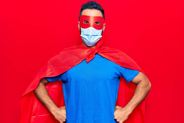 Junger hispanischer Mann in Superheldenkostüm und medizinischer Maske sieht positiv und glücklich stehend aus und lächelt mit einem selbstbewussten Lächeln, das Zähne zeigt  - Foto, Bild
