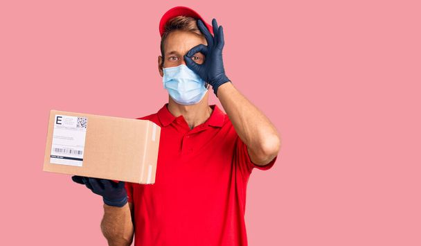 Handsome blond man with beard holding delivery box wearing medical mask smiling happy doing ok sign with hand on eye looking through fingers  - Photo, Image