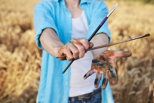 Close-up picture of dirty with paint hands, holding brushes, in front of yellow wheat field. Male artist wearing blue shirt and jeans painting in countryside. Outdoors artistic education activity. - Foto, Imagen