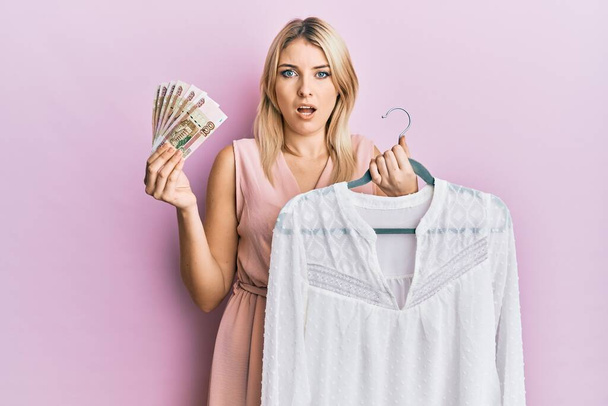 Young caucasian woman holding hanger with t shirt and russian ruble banknotes in shock face, looking skeptical and sarcastic, surprised with open mouth  - Фото, изображение