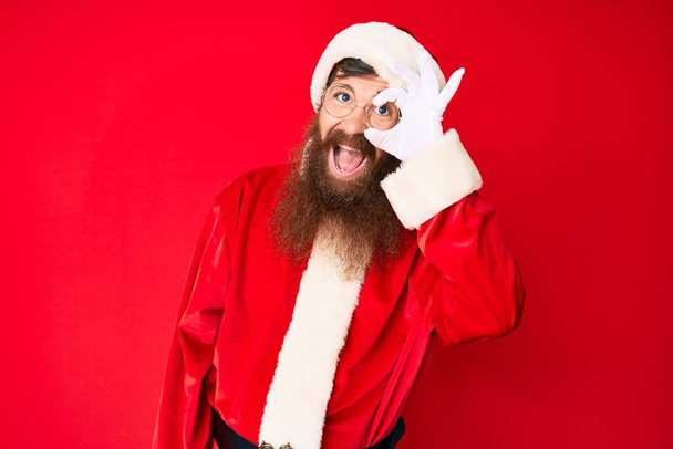 Handsome young red head man with long beard wearing santa claus costume smiling happy doing ok sign with hand on eye looking through fingers  - Zdjęcie, obraz