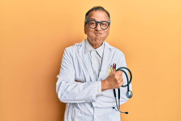 Indián středního věku v lékařském kabátě, držící stetoskop a čechrající tváře s legračním obličejem. ústa nadýchaná vzduchem, lapající vzduch.  - Fotografie, Obrázek