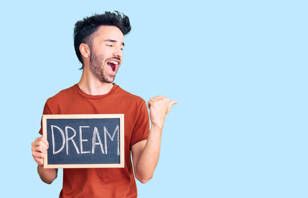 Young hispanic man holding blackboard with dream word pointing thumb up to the side smiling happy with open mouth  - Фото, изображение