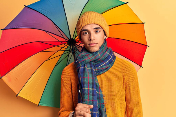Young african amercian man holding colorful umbrella thinking attitude and sober expression looking self confident  - Фото, изображение