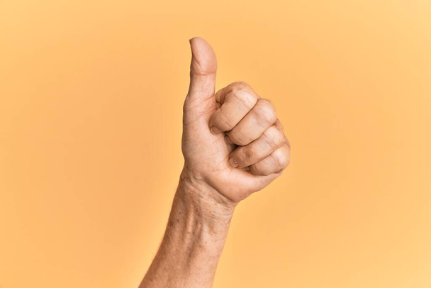 Senior caucásico mano sobre amarillo aislado fondo haciendo éxito aprobación gesto con pulgares hacia arriba, validación y símbolo positivo  - Foto, imagen
