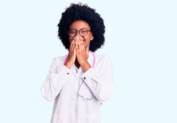 Молода афроамериканська жінка, одягнена в лікарське пальто і стетоскоп сміється і соромливо хихихикає, прикриваючи рот руками, плітками і скандалом концепції  - Фото, зображення