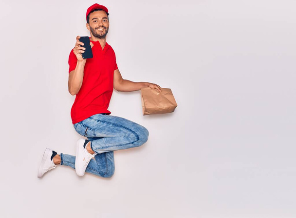 Young handsome hispanic deliveryman wearing uniform smiling happy. Jumping with smile on face holding paper bag with takeaway food and smartphone over isolated white background. - Photo, Image