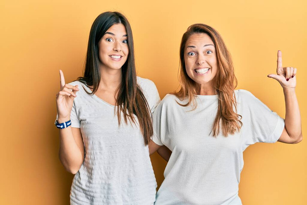 Latino perheen äiti ja tytär yllään rento valkoinen t paita hymyillen hämmästynyt ja yllättynyt ja osoittaa ylös sormilla ja nosti kädet.  - Valokuva, kuva