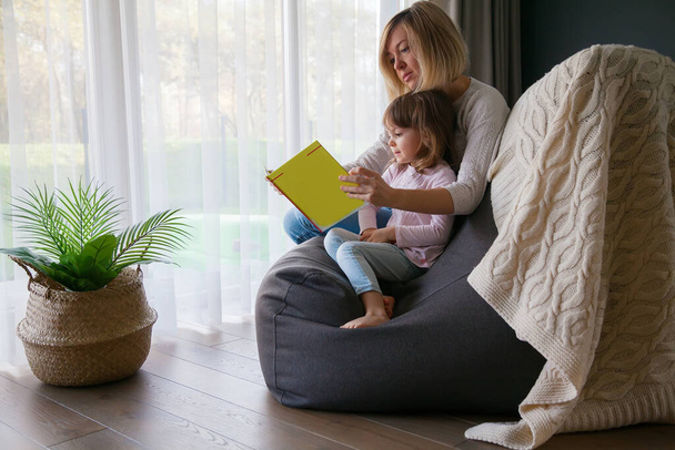 Mother reading a book to her little daughter, sitting on a bean bag chair at home - Photo, Image