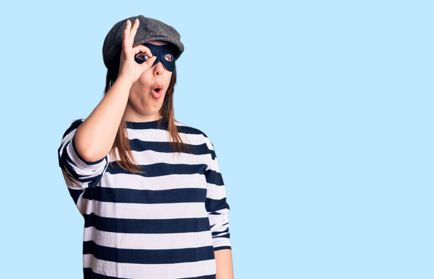 Young beautiful brunette woman wearing burglar mask doing ok gesture shocked with surprised face, eye looking through fingers. unbelieving expression.  - Photo, Image