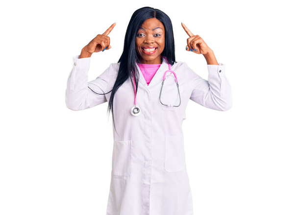 Young african american woman wearing doctor stethoscope smiling pointing to head with both hands finger, great idea or thought, good memory  - Fotografie, Obrázek