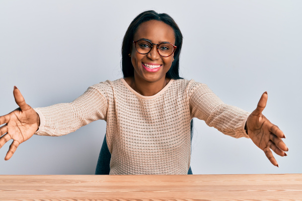 Eine junge Afrikanerin in legerer Kleidung sitzt auf dem Tisch und blickt lächelnd mit offenen Armen in die Kamera. fröhlicher Ausdruck, der das Glück umarmt.  - Foto, Bild
