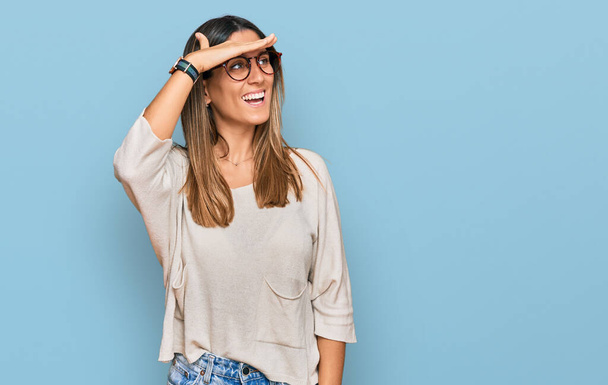 Mujer joven vistiendo ropa casual y gafas muy felices y sonrientes mirando lejos con la mano sobre la cabeza. concepto de búsqueda.  - Foto, imagen