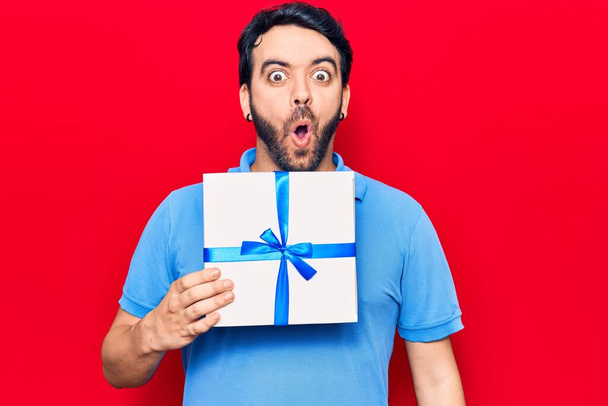 Young hispanic man holding gift scared and amazed with open mouth for surprise, disbelief face  - Fotoğraf, Görsel