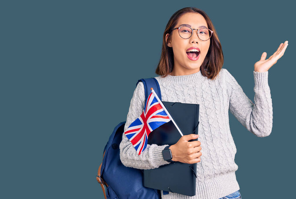 Fiatal gyönyörű kínai lány visel diák hátizsák gazdaság kötőanyag és Egyesült Királyság zászló ünneplő győzelem boldog mosollyal és a győztes kifejezés emelt kézzel  - Fotó, kép