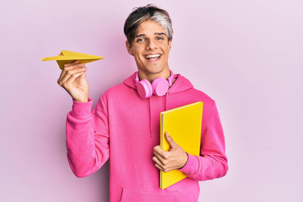 Νεαρός Ισπανός που κρατάει βιβλίο και χάρτινο αεροπλάνο χαμογελώντας με ένα χαρούμενο και δροσερό χαμόγελο στο πρόσωπο. που δείχνει δόντια.  - Φωτογραφία, εικόνα