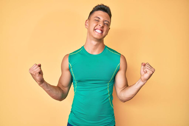 Fiatal spanyol fiú visel sportos stílus ujjatlan ing nagyon boldog és izgatott csinál győztes gesztus felemelt karokkal, mosolygós és sikoltozó siker. ünnepség koncepciója.  - Fotó, kép