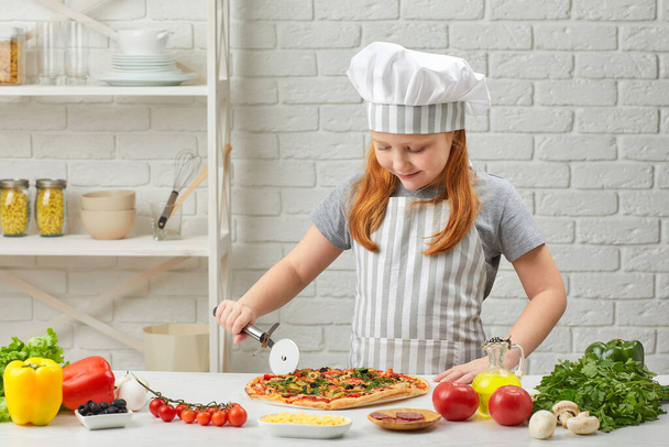cute little girl in chef hat and an apron cutting pizza - Фото, зображення