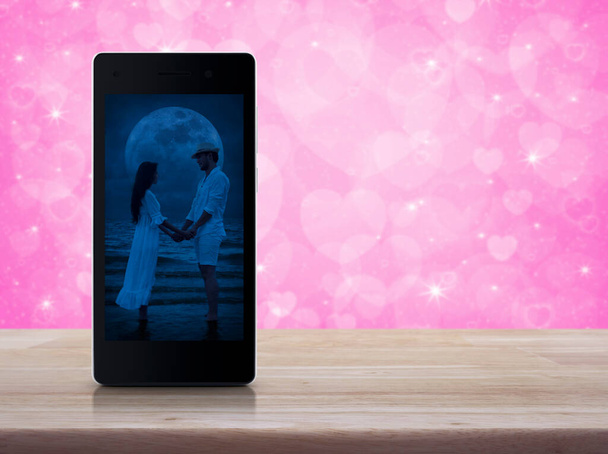 Romantic loving couple standing and holding hands together on modern smart mobile phone screen on wooden table over blur pink love heart background, Business internet dating online, Valentines day concept - Foto, Bild