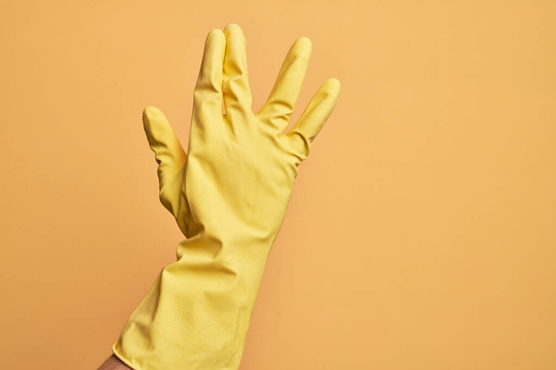 Mano de joven caucásico con guante de limpieza sobre fondo amarillo aislado saludo haciendo saludo vulcano, mostrando la parte posterior de la mano y los dedos, cultura freak - Foto, Imagen