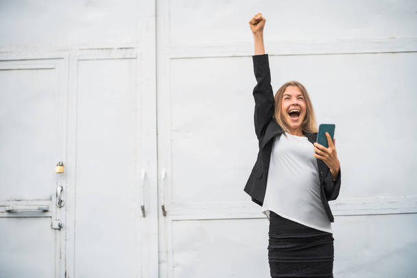 Außenporträt einer ekstatischen modernen Geschäftsfrau, die fröhlich ist. Sie hält ihr Smartphone in der Hand. - Foto, Bild