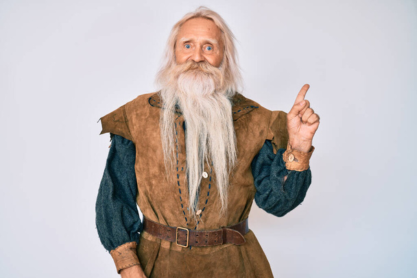 Vieil homme âgé aux cheveux gris et à la barbe longue portant un costume traditionnel viking avec un grand sourire sur le visage, pointant du doigt vers le côté en regardant la caméra.  - Photo, image