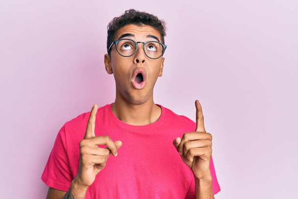 Молодий афроамериканець, одягнений в окуляри над рожевим фоном, вражений і здивований, дивлячись вгору і вказуючи пальцями і підняті руки.  - Фото, зображення
