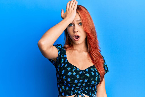 Jonge mooie roodharige vrouw draagt casual kleding over blauwe achtergrond verrast met de hand op het hoofd voor fout, herinner fout. vergeten, slecht geheugen concept.  - Foto, afbeelding