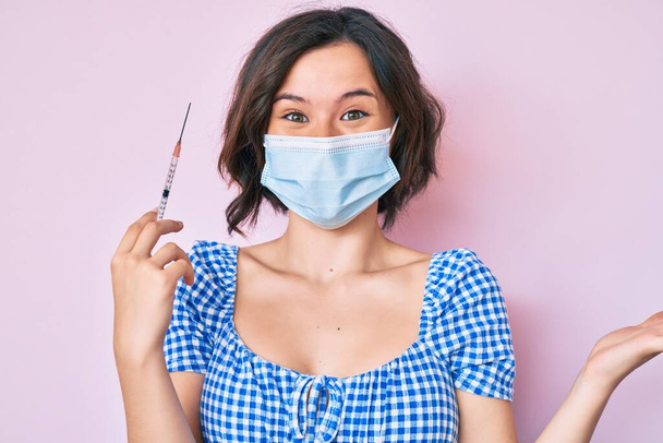Young beautiful woman wearing medical mask holding syringe celebrating victory with happy smile and winner expression with raised hands  - Photo, Image