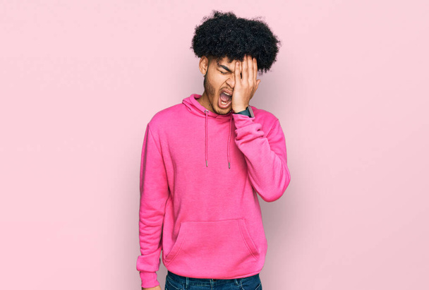 Ein junger afrikanisch-amerikanischer Mann mit Afro-Haaren trägt lässig ein rosafarbenes Sweatshirt, das das halbe Gesicht, Augen und Mund mit der Hand gähnt. Gesicht schmerzt vor Schmerzen.  - Foto, Bild