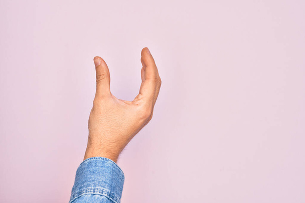 Dłoń białego młodzieńca pokazuje palce nad odizolowanym różowym tłem zbierając i biorąc niewidzialną rzecz, trzymając obiekt z palcami pokazującymi przestrzeń - Zdjęcie, obraz