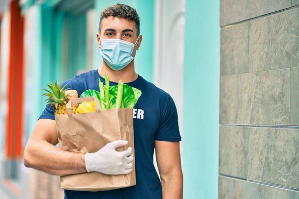Young hispanic volunteer man wearing medical mask holding groceries at the city. - Fotoğraf, Görsel