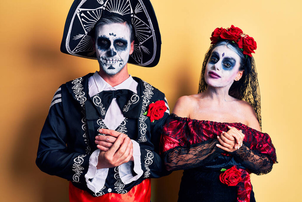 Jeune couple portant le jour mexicain du costume mort sur jaune avec les mains jointes et les doigts croisés souriant détendu et joyeux. succès et optimisme  - Photo, image