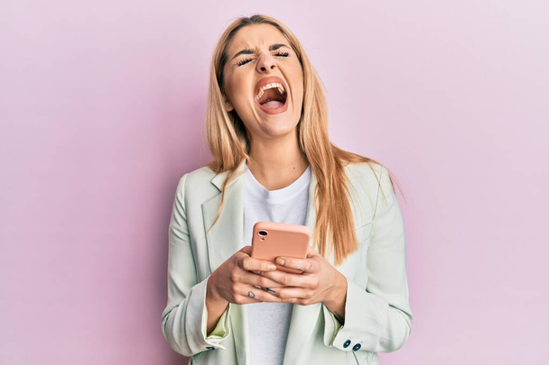 Jonge blanke vrouw draagt zakelijke kleding met behulp van smartphone boos en gek schreeuwen gefrustreerd en woedend, schreeuwen van woede opkijken.  - Foto, afbeelding