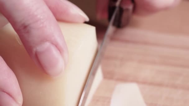 El queso de cabra prensado duro se corta en rodajas finas en una tabla de cortar de madera. Cocinar el desayuno europeo. Las manos femeninas cortan queso con un cuchillo. Enfoque selectivo. - Imágenes, Vídeo