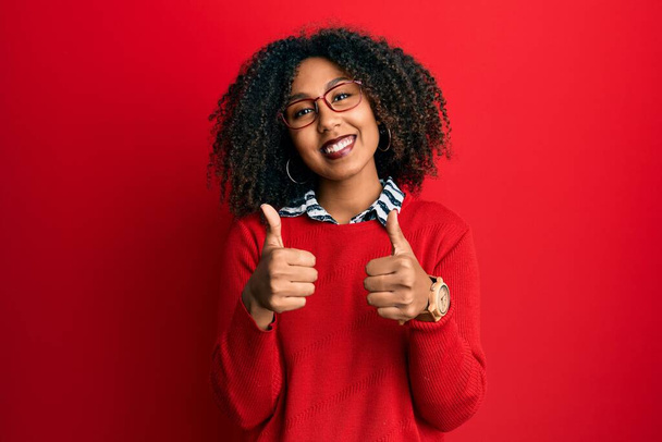 Bella donna afro-americana con i capelli afro indossa maglione e occhiali segno di successo facendo gesto positivo con la mano, pollice in su sorridente e felice. espressione allegra e gesto vincente.  - Foto, immagini