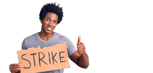 Нахабний афро-американець з волоссям афроамериканця, який тримає картонний плакат, посміхаючись щасливий і позитивний, великий палець робить відмінне і знак схвалення.  - Фото, зображення