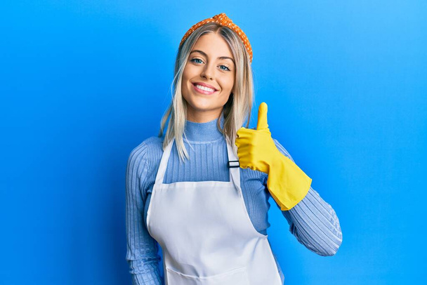 Beautiful blonde woman wearing cleaner apron and gloves doing happy thumbs up gesture with hand. approving expression looking at the camera showing success.  - Photo, image