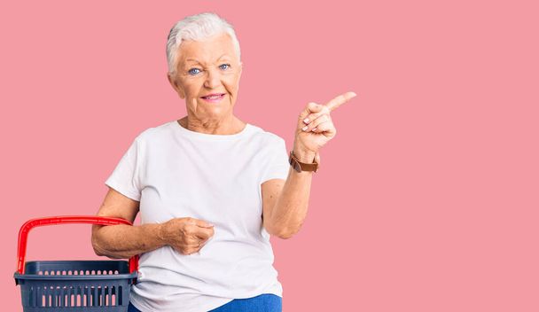Senior hermosa mujer con ojos azules y pelo gris sosteniendo cesta de la compra del supermercado sonriendo feliz señalando con la mano y el dedo a un lado  - Foto, Imagen