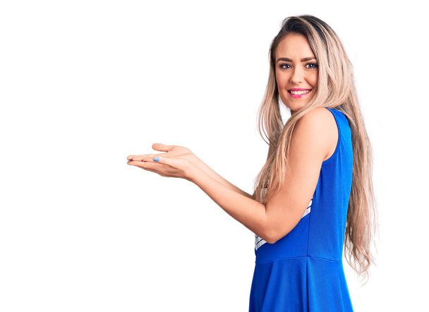 Молодая красивая блондинка в форме болельщицы указывая в сторону с открытыми руками ладонями показывая пространство для копирования, представляя рекламу улыбаясь взволнованный счастливый  - Фото, изображение