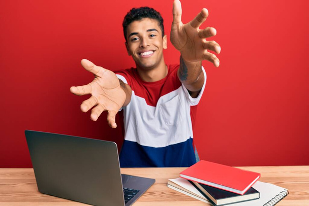 Jonge knappe Afro-Amerikaanse man studeert voor school met een laptop en kijkt naar de camera die glimlacht met open armen voor een knuffel. vrolijke uitdrukking omarmen geluk.  - Foto, afbeelding