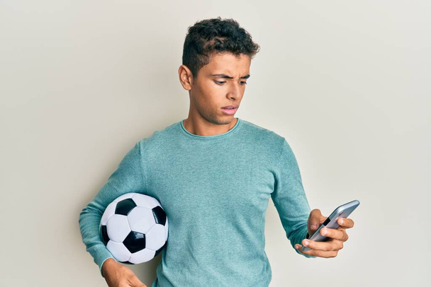 サッカーボールを持っている若いハンサムなアフリカ系アメリカ人男性は、スマートフォンの無知で混乱した表情を見ています。疑わしい概念.  - 写真・画像
